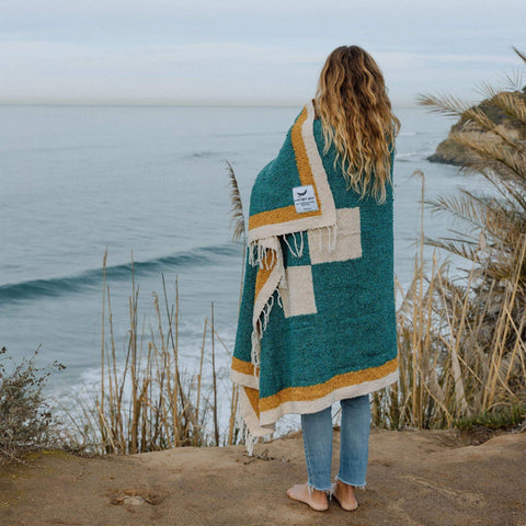 Person wrapped in a Trek Light Dolly Blanket overlooking the ocean, showcasing eco-friendly, handwoven design.