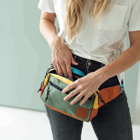 Woman wearing Everyday Fanny Pack (Cypress) by Trek Light, convertible from hip pack to shoulder sling, in vibrant colors.