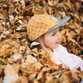 Child wearing a yellow Flower Child Kids Hat by Trek Light, lying in autumn leaves, showcasing the stylish 5-panel snapback design.