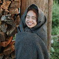 Woman joyfully wrapped in a cozy Trek Light Greyson Blanket, handwoven from recycled clothing materials, showcasing its versatile beauty.