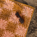 Groovy Flowers Bath Towel with retro floral design in pink and orange, featuring Trek Light's sustainable cotton, laid on sandy beach.