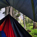 Trek Light V9 Strong Tarp with hammock in forest setting under a colorful rainbow sky.