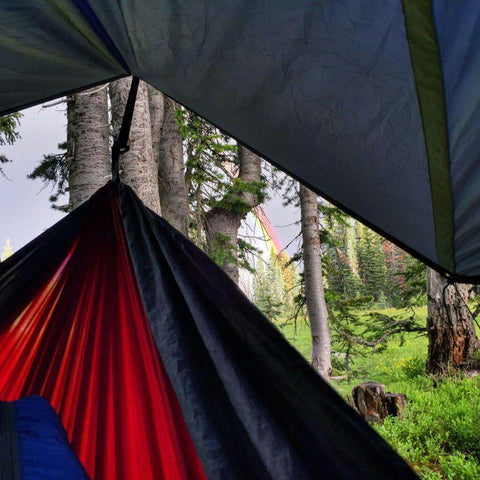 Trek Light V9 Strong Tarp with hammock in forest setting under a colorful rainbow sky.
