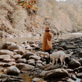 Woman by a river with dog, carrying Trek Light leather blanket carrier through nature's rocky terrain.