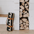 Stylish Trek Light leather blanket carrier holding a cozy patterned blanket against a wood pile backdrop.