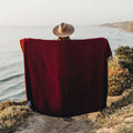 Person wrapped in red Napa Blanket by the ocean, cozy and eco-friendly, perfect for naps or enjoying nature. Trek Light.