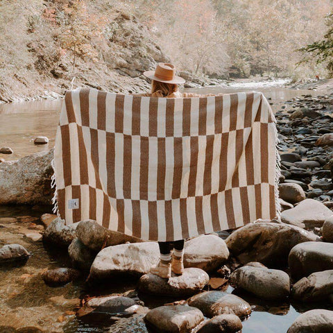 Person holding a Trek Light striped blanket by a serene river, surrounded by nature during a sunny day.