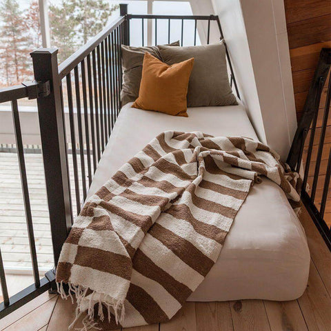 Striped brown and white blanket draped over bed with pillows, cozy setting by windowsill. Perfect addition to home decor.