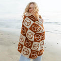 Cozy woman wrapped in Trek Light blanket with peace symbols, smiling by the beach.