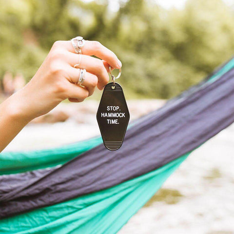 Hand holding a black "Stop. Hammock Time." key tag with green hammock in background. Perfect for a relaxing lifestyle. Trek Light.