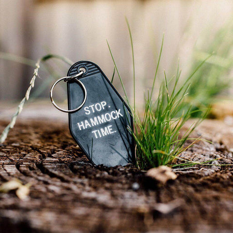 Black "Stop: Hammock Time" key tag on a wooden stump with greenery, perfect for a relaxing vibe. Keywords: Trek Light, lifestyle accessory.