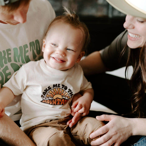 Happy child in 'Take Me To The Mountains' Trek Light shirt, surrounded by parents, promoting outdoor play and adventure.