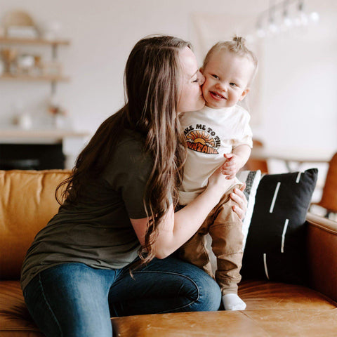 Mom hugging happy toddler wearing 'Take Me To The Mountains' Trek Light shirt, perfect for outdoor play and family adventures.