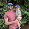 Father and child wearing matching 'Take Me To The River' Trek Light hats in front of evergreen trees, enjoying a sunny outdoor day.