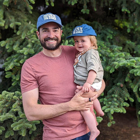 Father and child wearing matching 'Take Me To The River' Trek Light hats in front of evergreen trees, enjoying a sunny outdoor day.