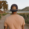 Man wearing 'This Must Be The Place' mountains hat by Trek Light, enjoying scenic outdoor view, stylish snapback adventure gear.