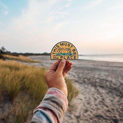 Hand holding "This Must Be The Place" beach sticker by Trek Light, featuring palm trees and sunset, against a serene beach background.